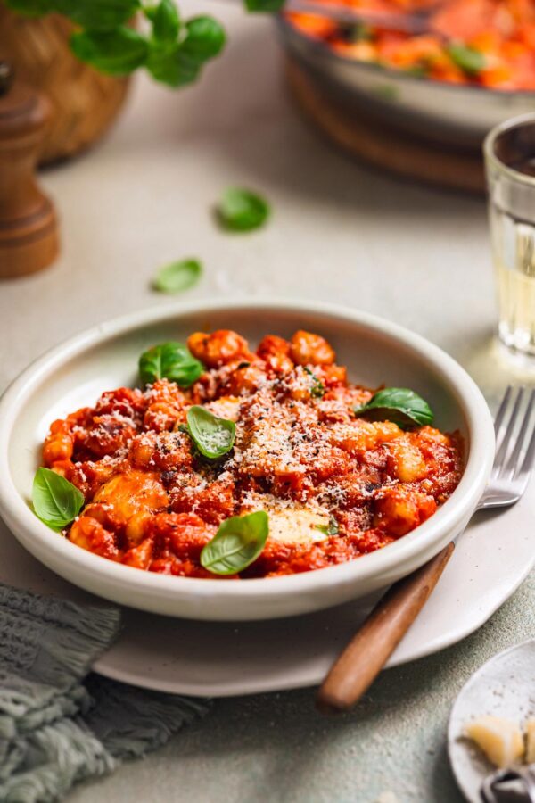 Ein Teller Gnocchi in Tomatensauce, überzogen mit Parmesan und verziert mit Basilikumblättern. Eine Gabel liegt griffbereit am Tellerrand.